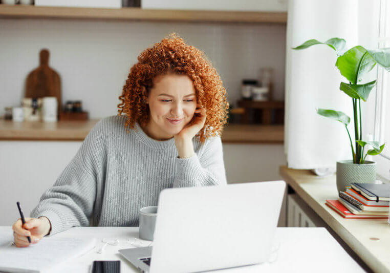 Cute girl with red curls watching online tutorial or lesson on laptop, smiling listening and looking attentively, making notes in copybook, sitting at kitchen table with smartphone and cup of coffee
